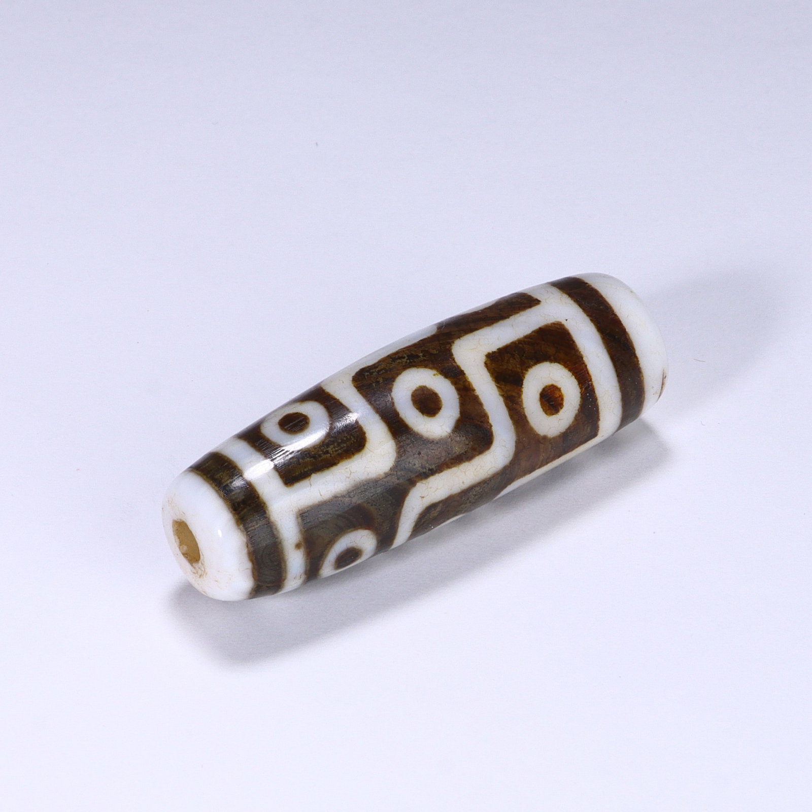 CHINESE NINE-EYE DZI BEAD: Dimensions:L. 4 cm. A Chinese cylindrical Dzi bead, measuring 4 cm in length, featuring a creamy ivory base with rich brown geometric patterns. The bead displays nine circular "eye" motifs arranged in