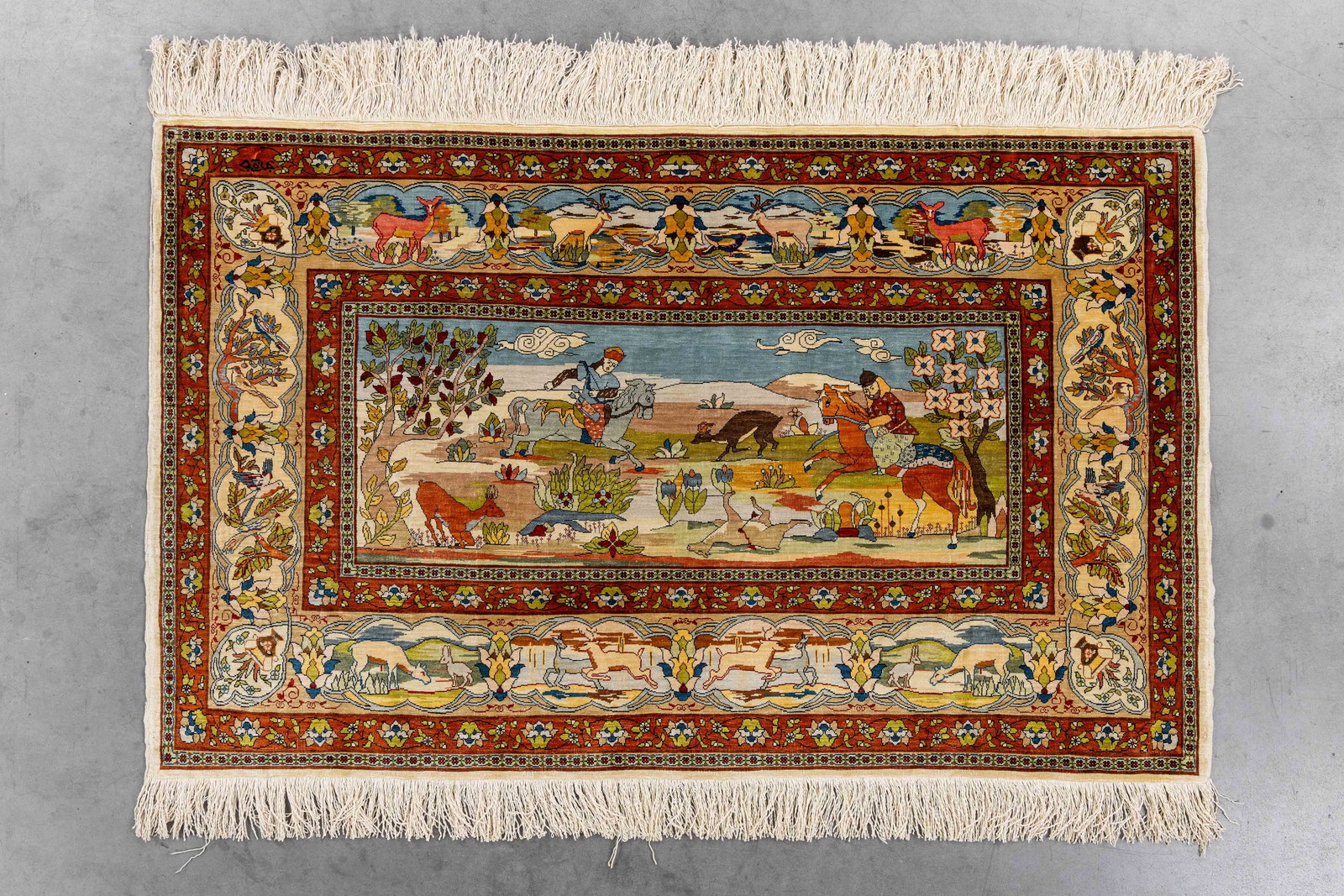 An Oriental hand-made carpet, Silk, Hereke, Turkiye (112 x 69 cm).: An Oriental hand-made carpet, Silk, Hereke, Turkiye (112 x 69 cm). With figurative hunting decor. Signed. (69 x 112 x 0 cm)
