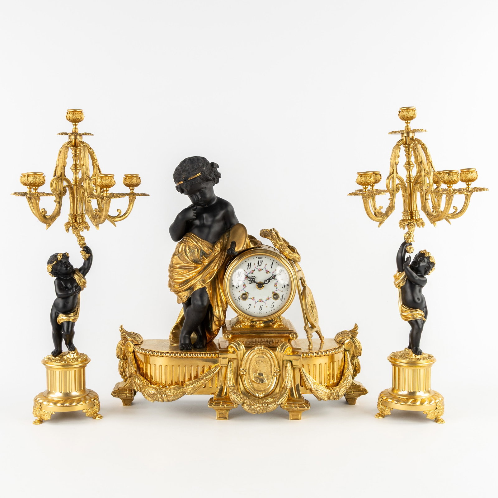 A three-piece mantel garniture clock and candelabra, gilt and patinated bronze, finished with putti.: A three-piece mantel garniture clock and candelabra, gilt and patinated bronze, finished with putti. 20th century. The clock finished with a putti, gilt and patinated bronze. Putti with a painter's pa