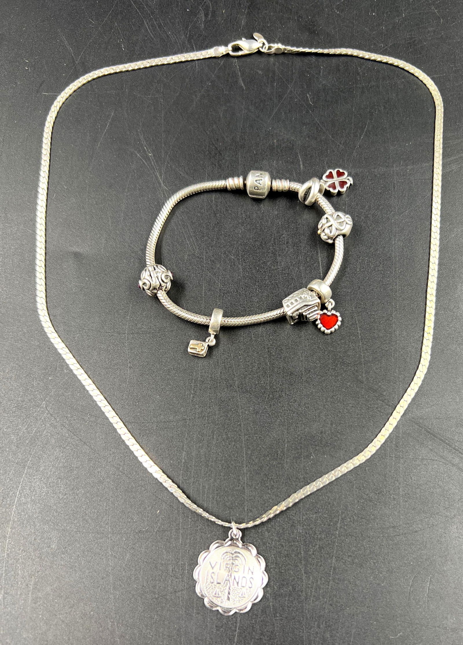 Sterling Silver Virgin Islands Necklace and Sterling Silver Pandora Red Charm Bracelet: In this lot is a Sterling Silver Virgin Island pendant with a Sterling Silver Herringbone Chain alongside a Pandora Sterling Silver charm bracelet with red charms. The necklace weight is 0.36 ozt with