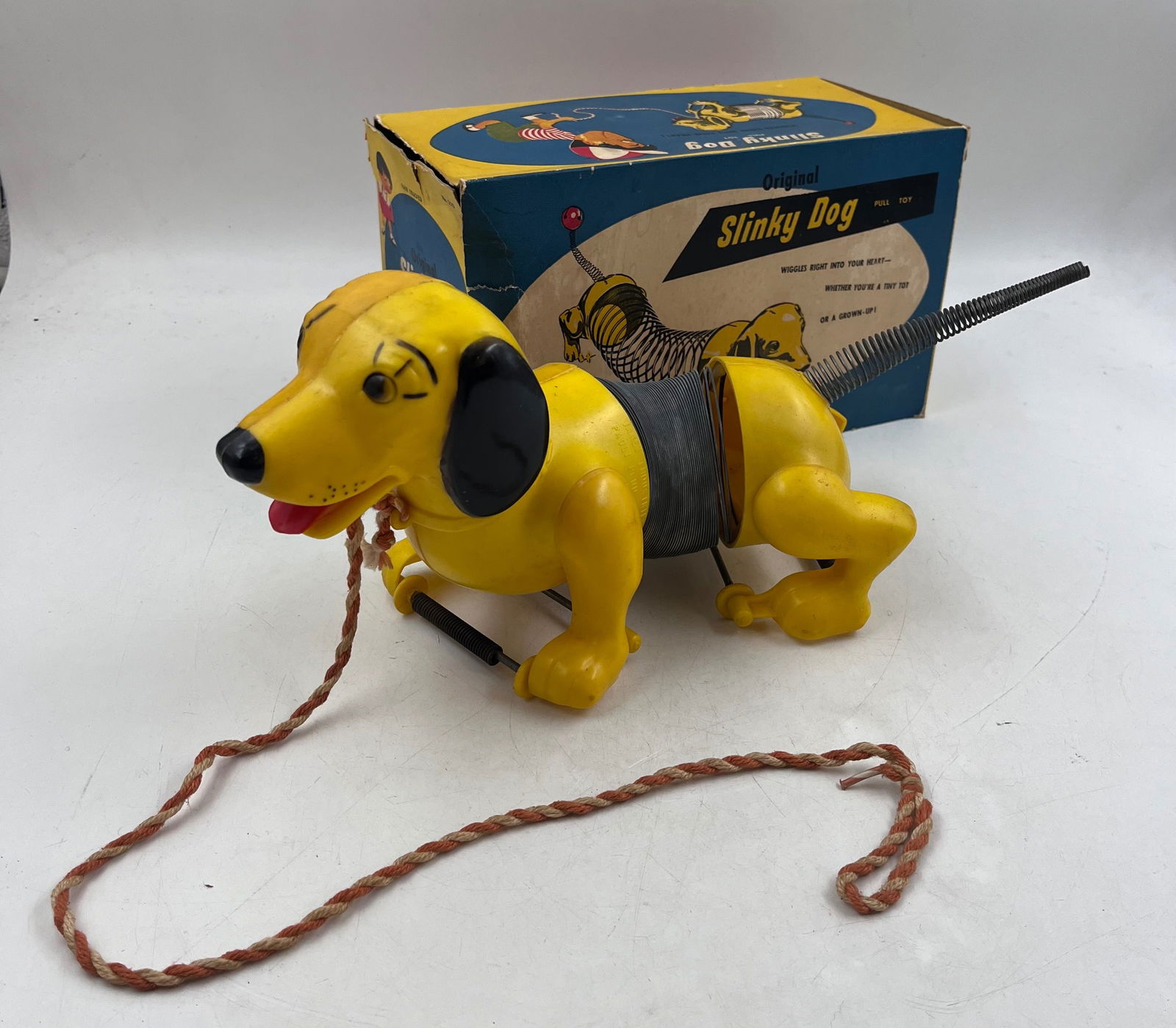 1950s Slinky Dog Pull Toy in Original Box: 1950s Slinky Dog Pull Toy in Original Box. The slinky dog was made by James Industries. The toy is of a yellow dog with a slinky body. A rope is attached so the toy can be pulled. The dog has a smalle