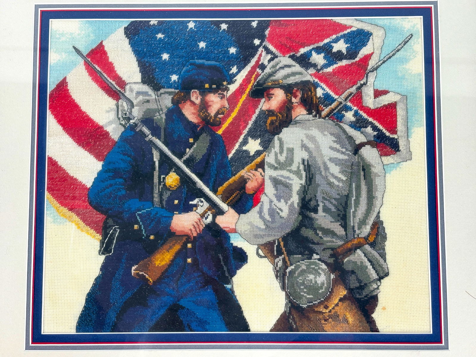 Cross Stitched Soldiers: Crossed stitch image of two soldiers facing off with one another wearing Confederate and Union Uniforms, with the respective flags in the background. Frame measures 20.5in x 18.25in