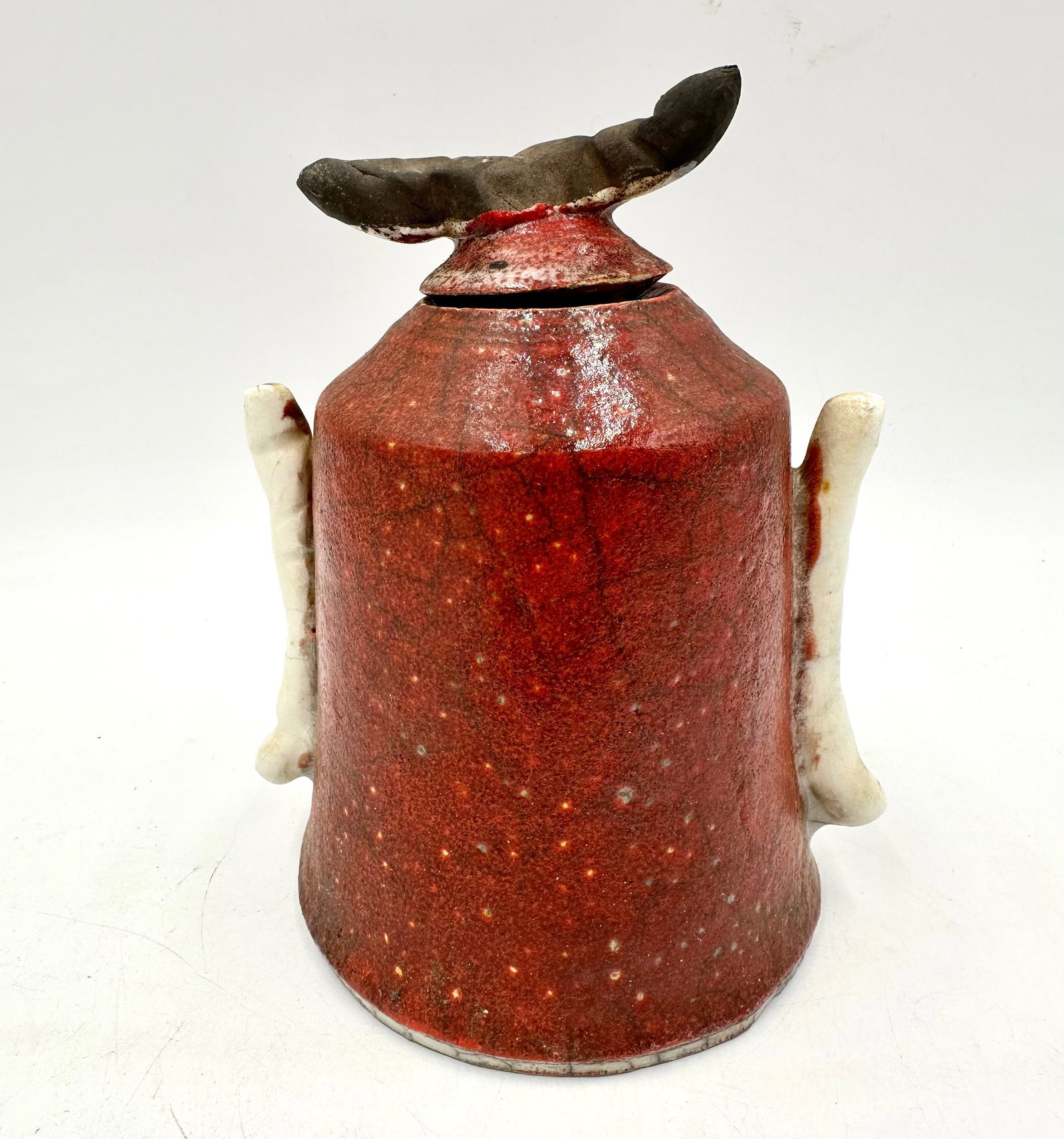 Han Red Raku Jar with Lid: The Ceramic jar with a lid has been done in a raku style. The red glaze shows a crackle design from the fire. The lid has a coiled handle. The jar has two coiled handles that match the lids handle alt