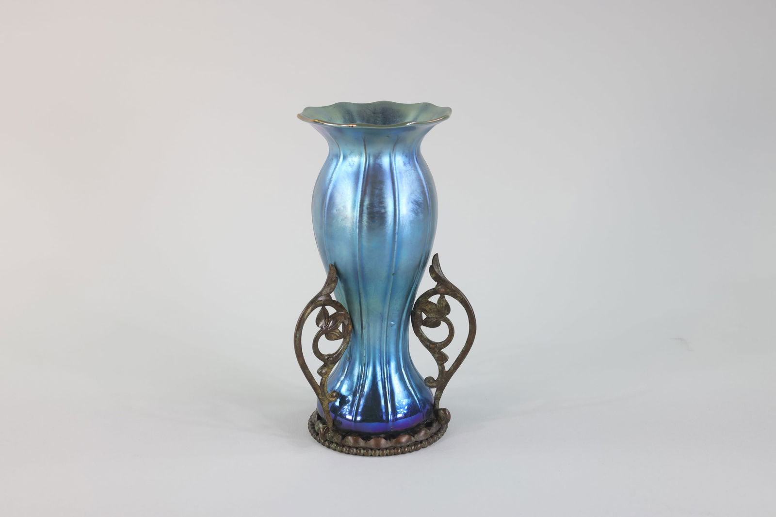 Tiffany Studio Favrile Glass with Bronze Base: A Tiffany Studio blue metallic Favrile glass with an Art Nouveau-style bronze base. The vase has a curving body and line work texture like a flower petal. The bronze base is flat with curvin