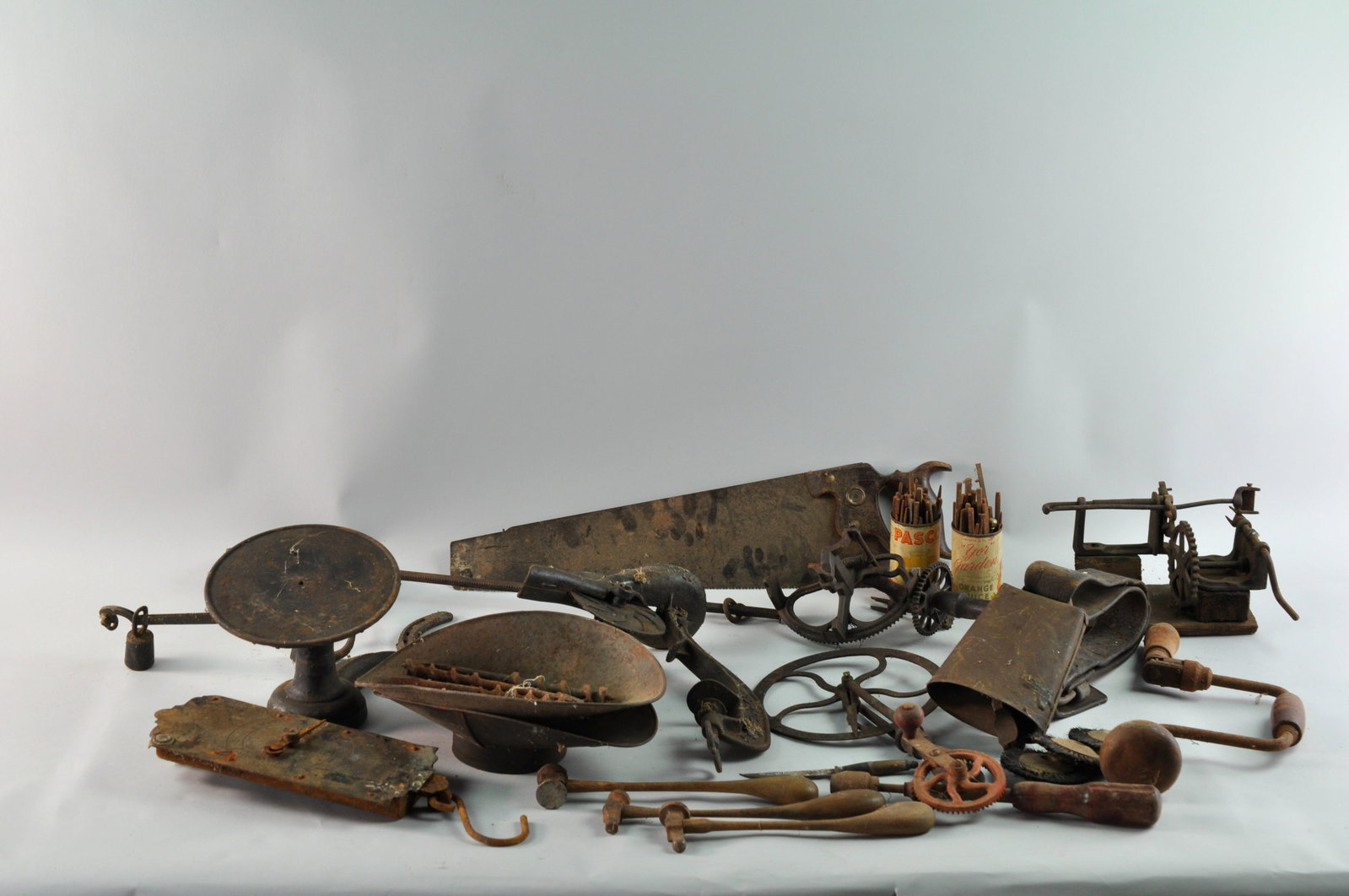 Cast Antique Scales and Tools: An assortment of antique tools, including a milk scale, a warranted saw with a wood handle, part of a cast iron butcher scale with three hooks and a star, a cowbell on a leather strap, two chasing ham