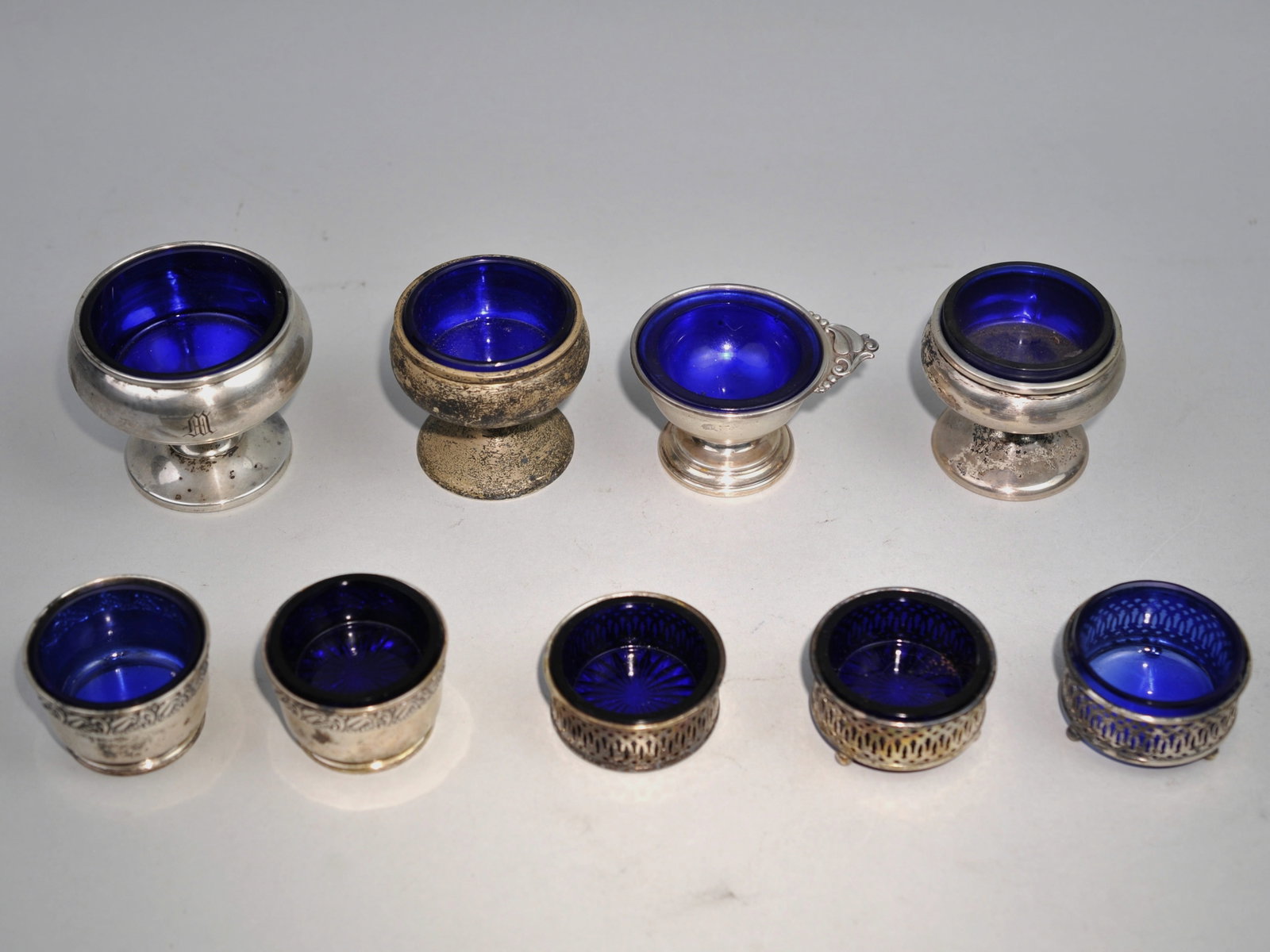 Sterling Silver Salt Cellars w/Blue Inserts: Metal: Sterling Silver Manufacturer:?Various Dimensions:?Various Condition: The Silver has Patina Additional Information:?This striking collection of nine sterling silver open salt cellars commands
