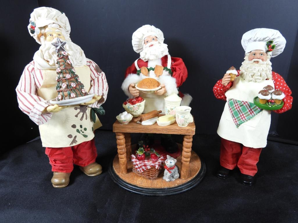 LOT OF 3 FABRIC Chef Baking Santas: LOT OF 3 FABRIC Chef Baking Santas: 11"T Santa baking pies at table of baking items and gray cat on circular 7" wide diameter platform, Chef Santa with spoon behind his ear and chocolate tree cake on