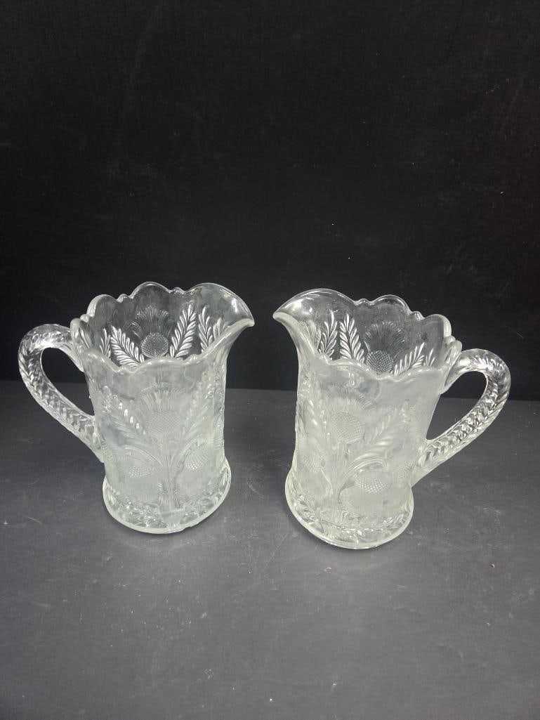 Pair of Pressed Glass Wheat and Thistle Pattern Pitchers: Matching pair of tall cylindrical pitchers, wheat sheaf and thistle floral panel pattern, pinched spout, applied handle, scalloped rim. Approx. 6.5"H x 4.5"W each.