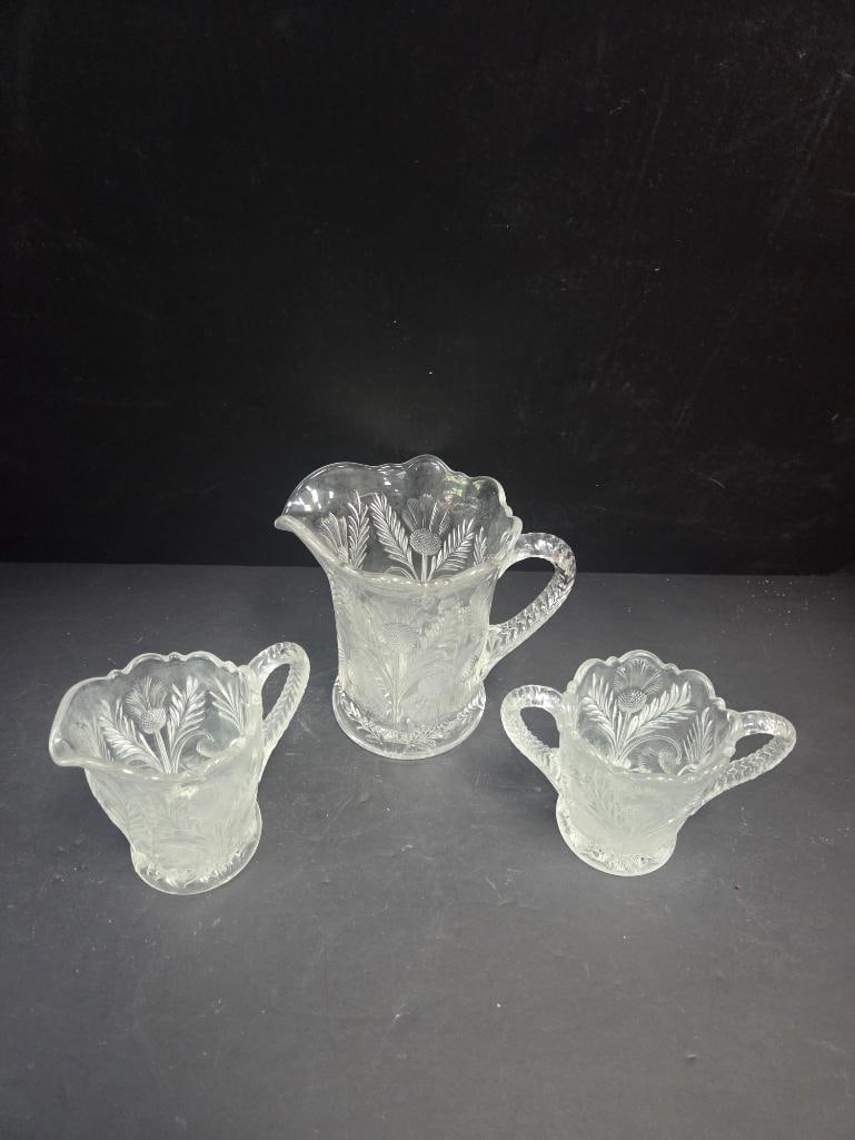 Three-Piece Pressed Glass Thistle Pattern Table Set: Pitcher, thistle pattern body, pinched spout, braided applied handle, scalloped rim. Approx. 6"H x 4.5"W.Small creamer, matching thistle pattern and braided handle. Approx. 3.5"H x 3.5"W.Open sugar bo