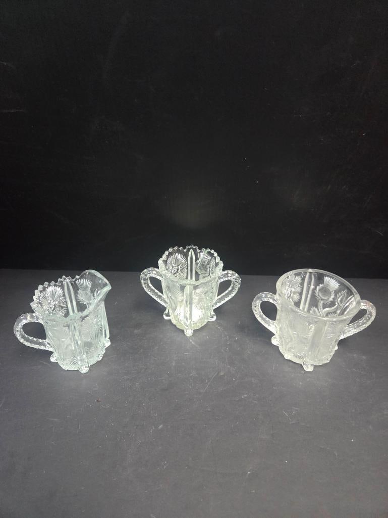 Three-Piece Pressed Glass Thistle Pattern Table Set, Footed: Small creamer, thistle pattern body, single applied handle, footed. Approx. 3.5"H x 3.5"W.Open sugar bowl, matching thistle pattern, two applied handles, footed. Approx. 3.5"H x 4"W.Small creamer,