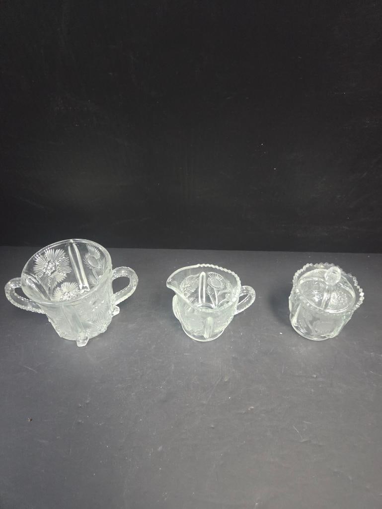 Mixed Grouping of Pressed Glass Sugar and Creamers: Open sugar bowl, starburst pattern body, two applied handles, footed. Approx. 3.5"H x 4"W.Creamer, floral/thistle pattern body, single applied handle, pinched spout, footed. Approx. 3.5"H x