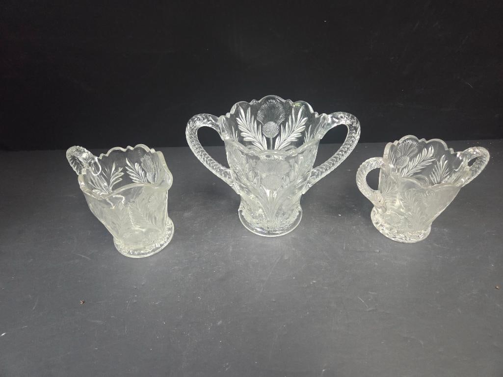 Three-Piece Pressed Glass Fern Pattern Table Set: Small creamer, fern/leaf etched pattern body, single applied handle, footed. Approx. 3.5"H x 3.5"W.Open sugar bowl, matching fern/leaf pattern, two applied handles, footed, scalloped rim (taller than