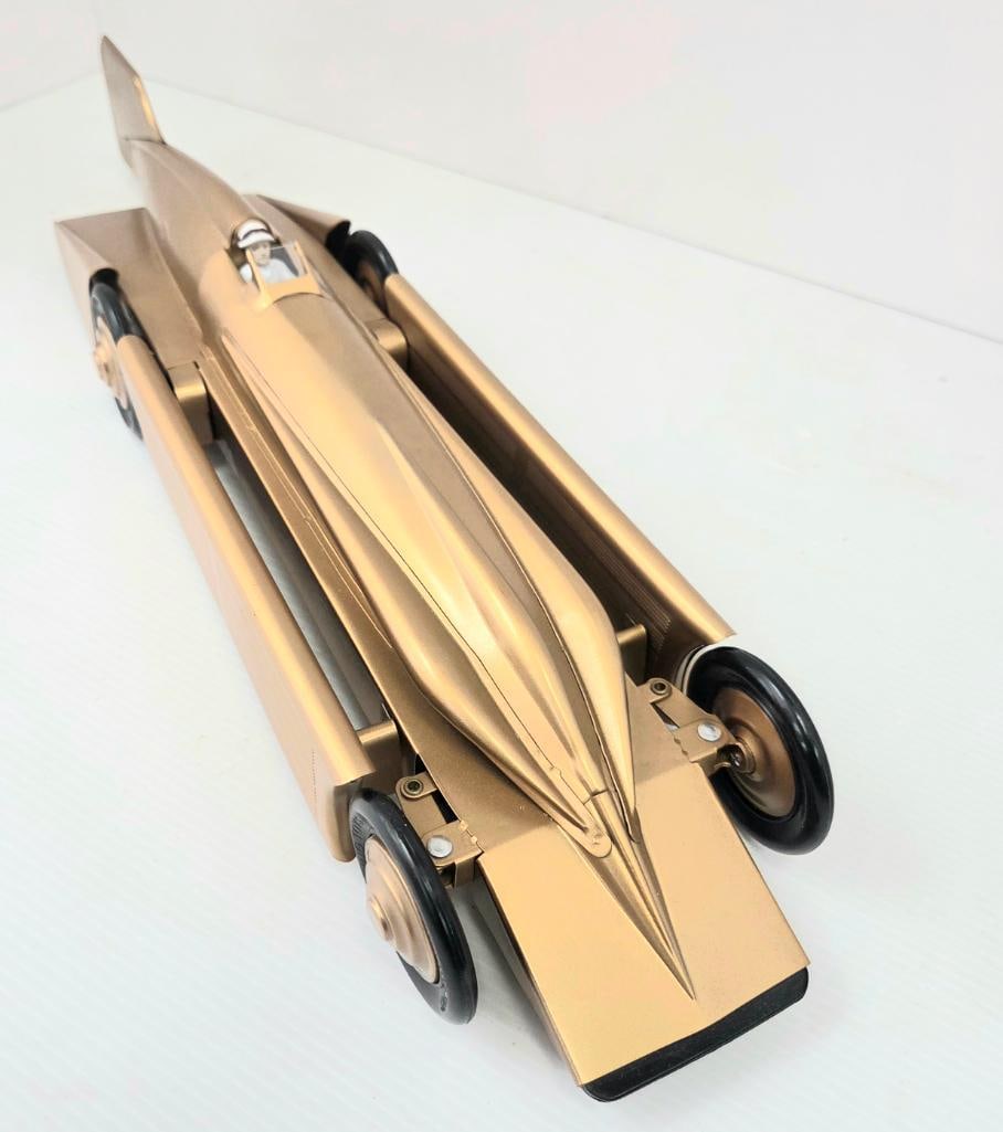 Gold-Tone Land-Speed-Record Replica Race Car: Gold/bronze-finished tin or metal replica land-speed-record style race car, driver figure in helmet, tail fin, streamlined body similar in style to the Bluebird replica in Lot 247 but in a different