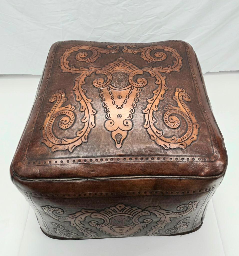 Tooled Leather Box/Ottoman with Scrollwork Design: Tooled leather-covered box or footstool/ottoman with a shaped, slightly domed lid, embossed scrolling acanthus-leaf and shield motif on the top and each side panel, brass-studded trim along the edges,