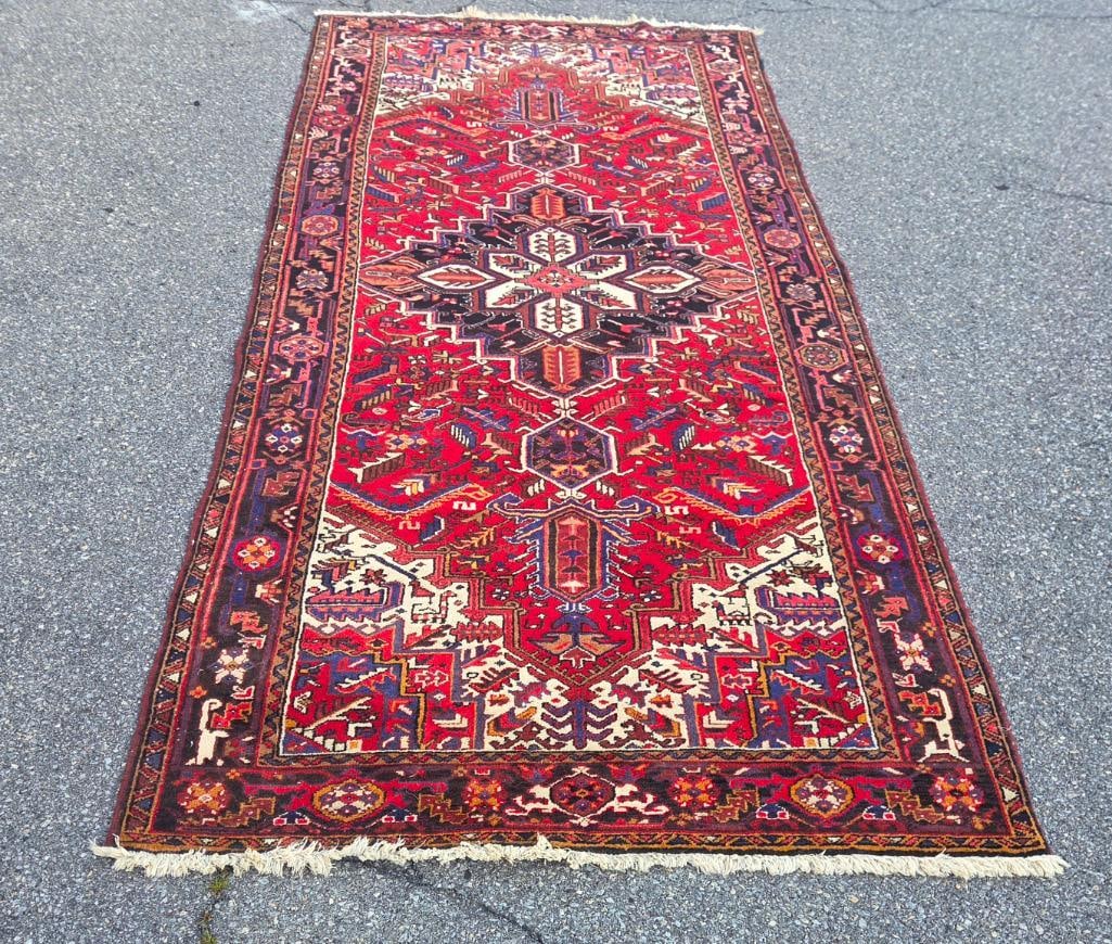 Red Heriz-Style Persian Wool Rug: Hand-knotted red Heriz-style Persian wool rug, large geometric star medallion in ivory, navy, and gold, angular floral field motifs, dark indigo border, fringed ends, approx. 5' x 9'. No maker's label