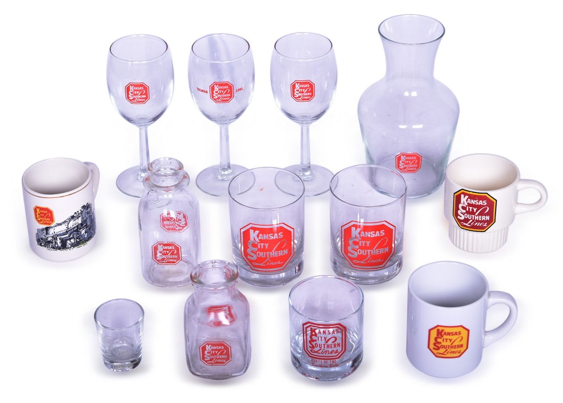 Thirteen Pieces of Kansas City Southern Railroad Glassware - Carafe, Wine Glasses, Shot Glass, Milk: A mixed group of drinkware all marked for Kansas City Southern Lines Railroad with enamel along the side. Includes 8" tall glass carafe, three 7" stemmed wine glasses (one marked Tolmak 1991), two 4" 