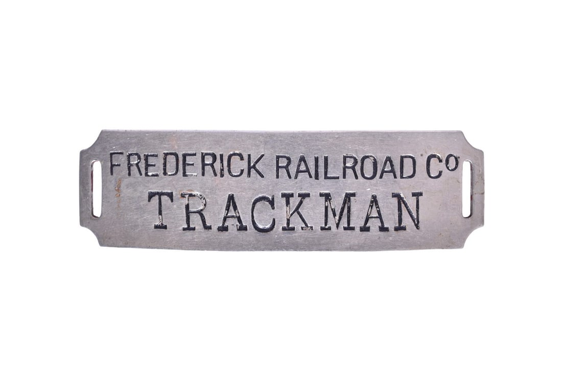 Frederick Railroad Co. Trackman Nickel Cap Badge: A 3 1/8" by 1" nickel plated Trackman cap badge with black enamel lettering and slot at each end. Shows general wear with discoloration and some chips throughout the enamel. The Frederick Railroad Co.