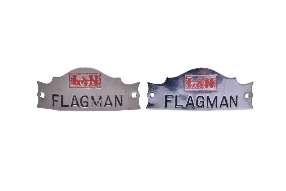 Pair of Louisville and Nashville Railroad Flagman Cap Badges: Two flagman badges of different metals each with the L&N logo in red enamel above the 'Flagman' title. Both badges are roughly 3 3/4" long by 1 1/2" tall, and have holes at each end for brads. One of 