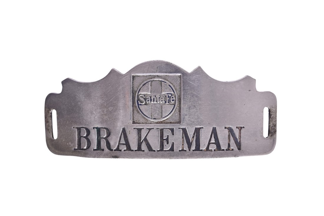 A nickel Brakeman cap badge for the Santa Fe Railway, stamped with the Santa Fe logo, showing light