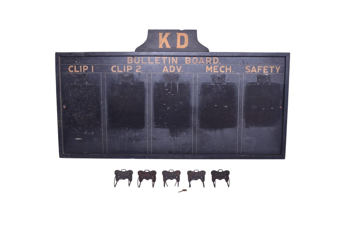 56" by 26" bulletin board with a smaller 17 1/2" by 5 1/4" board painted K D. The board has 5: 56" by 26" bulletin board with a smaller 17 1/2" by 5 1/4" board painted K D. The board has 5 spaces for hanging clipboards, labelled Clip 1, Clip 2, Adv., Mech., and Safety. The hardware for hanging 