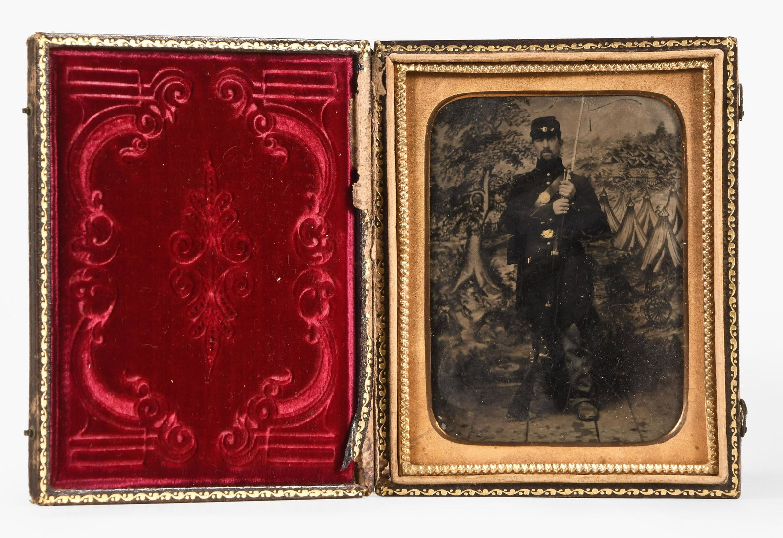 A good cased quarter plate tintype featuring a Union soldier in full uniform with his rifle: A good cased quarter plate tintype featuring a Union soldier in full uniform with his rifle, standing before a canvas backdrop depicting an encampment, with tents and cannon, contained in an embossed,