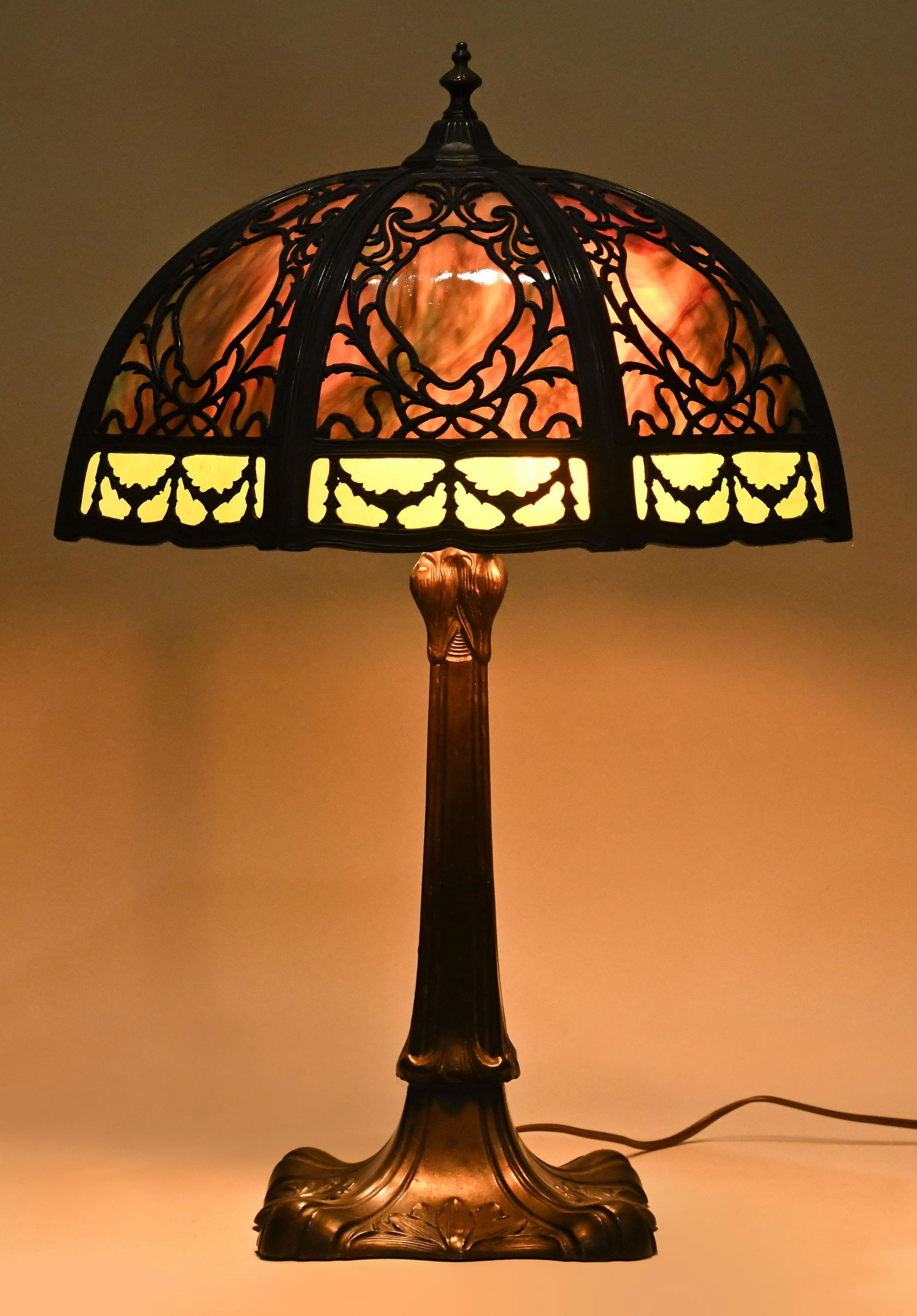 A good early 20th century Miller style metal overlay table lamp: A good early 20th century Miller style metal overlay table lamp, domed octagonal two color slag glass shade, the bronze finished frame with stylized leaf, bowknot, and swag ornament, and bronze finish