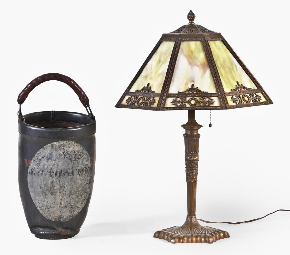 An early 20th century slag glass table lamp and a 19th century fire bucket: An early 20th century cast iron table lamp, fluted and gadrooned hexagonal base in a bronze finish and supporting a tapered column with acanthus and Vitruvian wave ornament below a cylindrical reeded 