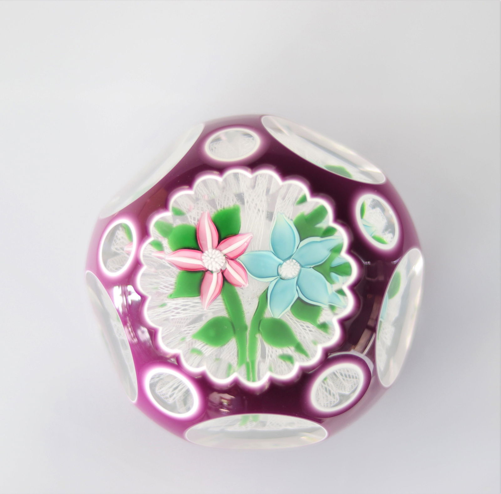 John Deacons 2000 paperweight, white and mauve double overlay with pink and blue flower on green: John Deacons 2000 paperweight, white and mauve double overlay with pink and blue flower on green stem on filagram cushion - Weight: 515 g - Sizes: H=55mm D=80mm - Condition: good condition - Author /