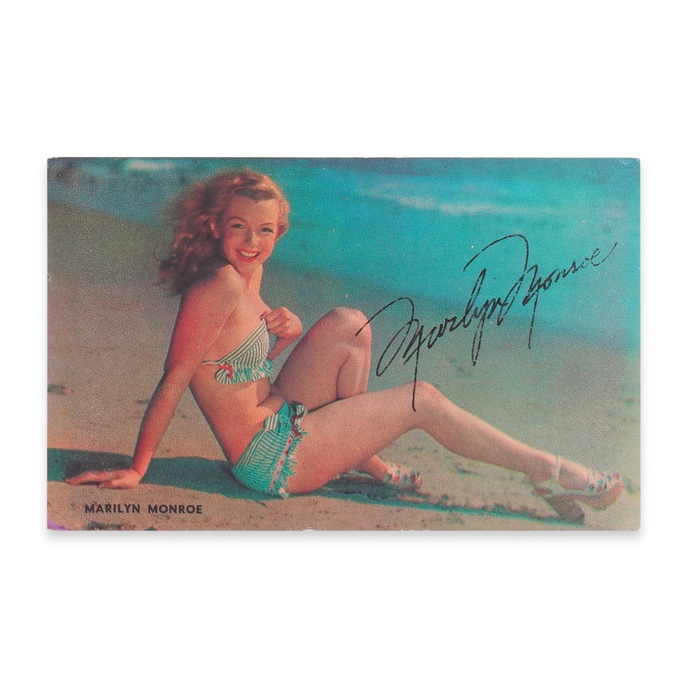 MARILYN MONROE SIGNED POSTCARD - BEACH POSE: 5 1/2 x 3 1/2 in (14 x 9 cm) This signed postcard featuring Marilyn Monroe captures the essence of Hollywood glamour at its most iconic moment. The image, depicting Monroe in a relaxed beach pose,