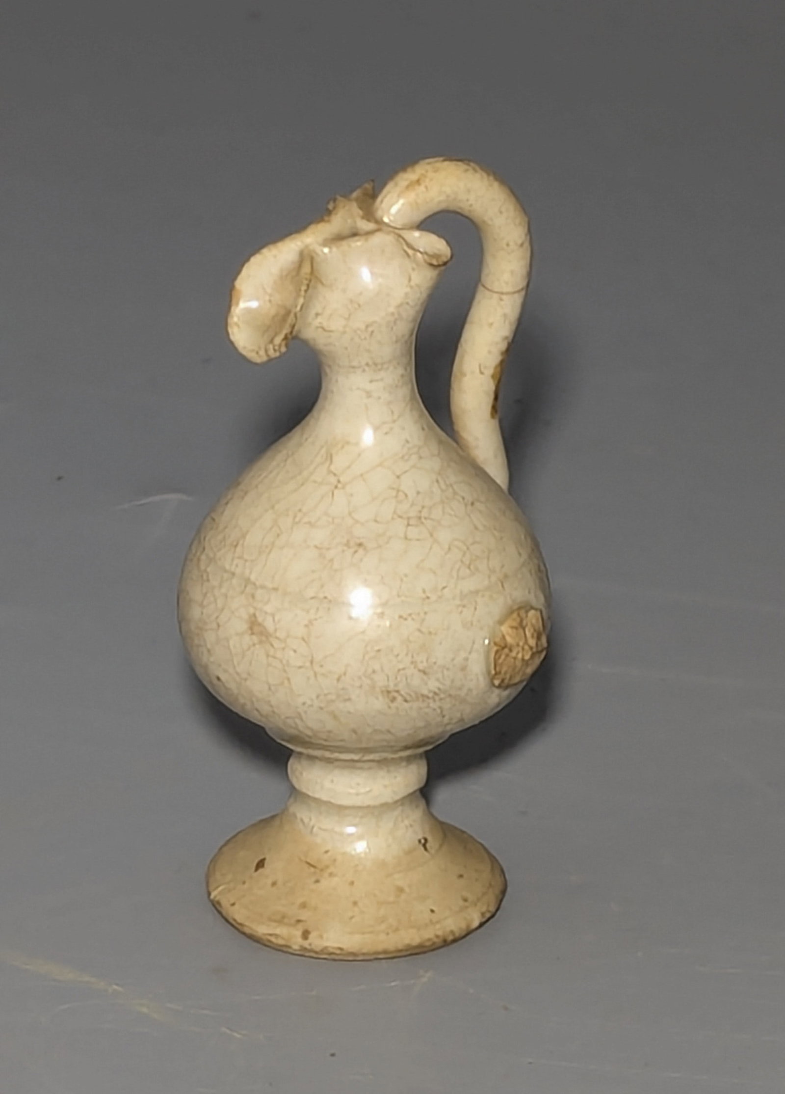 Chinese White-Glazed Miniature Ewer, Tang / Liao Dynasty Style: A small Chinese ceramic ewer, finely potted with a bulbous body raised on a spreading hollow pedestal foot, the tapering neck terminating in a stylized pinched, bird-like spout. A high arched handle e