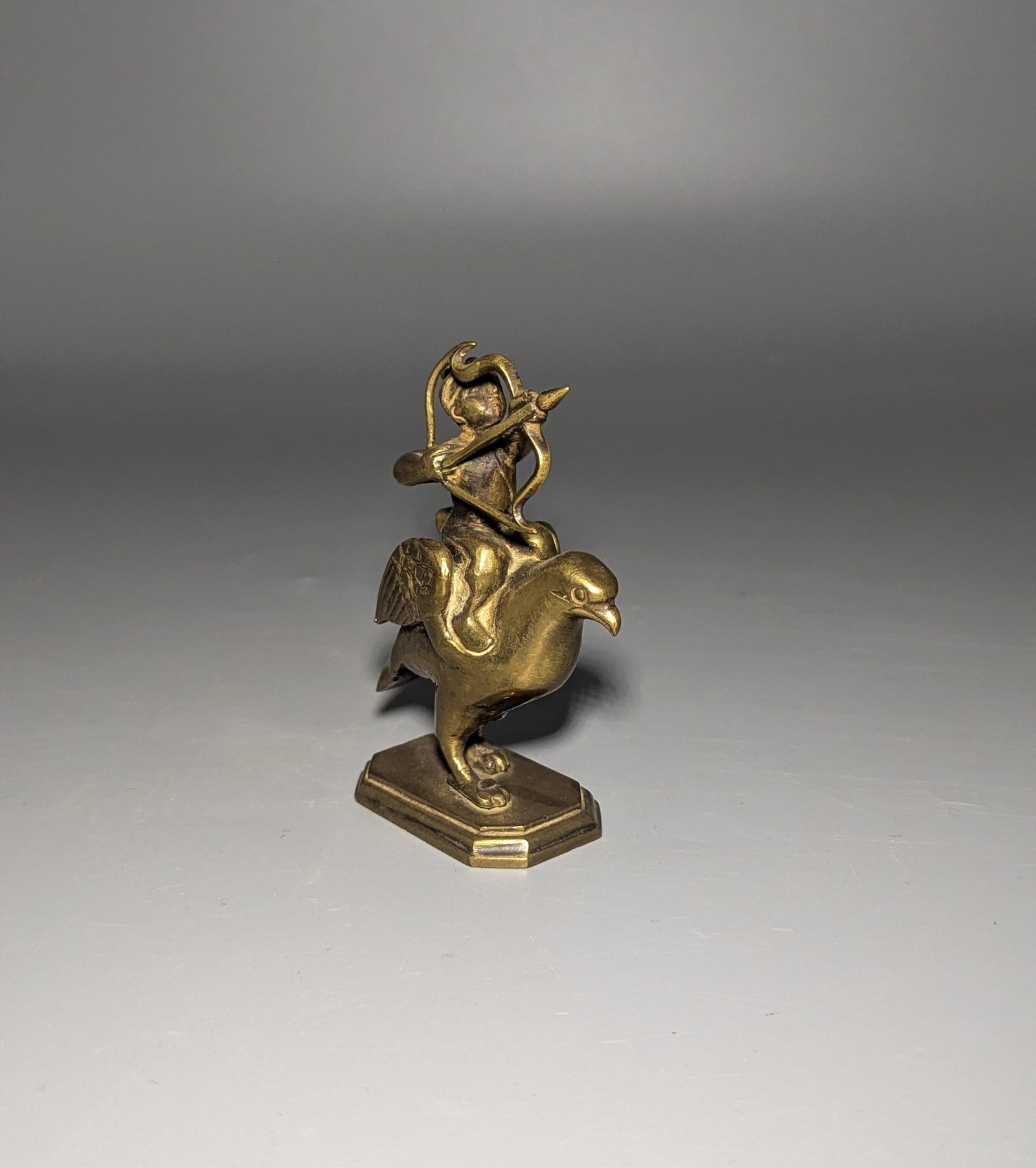 Indian Brass Figure of Kamadeva on a Bird Vahana: A small Indian cast brass devotional figure, probably representing Kamadeva, the Hindu deity of love and desire, modeled as an archer drawing his bow while seated upon an avian vahana. The figure wear