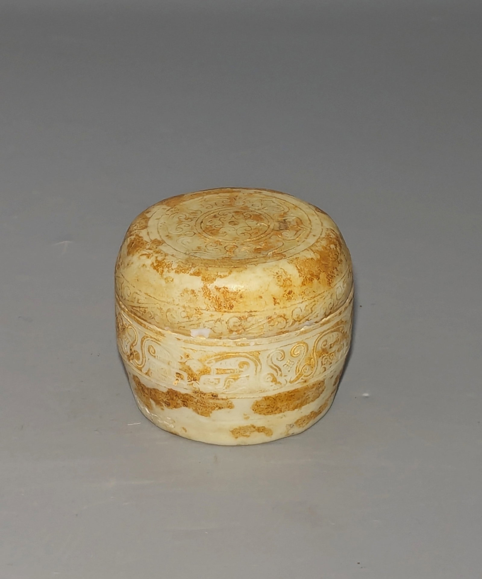 Chinese Archaistic Carved White and Russet Jade Seal Paste Box and Cover: Of circular cylindrical form, the fitted cover and lower container are carved in low relief, the cover centered by a stylized floral medallion surrounded by scrolling ruyi-form motifs, with the sides 