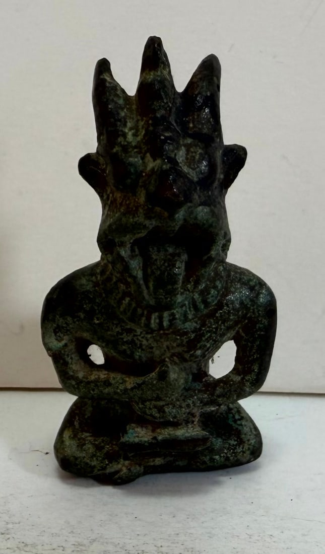 Bronze Seated Three Horned Deity: Apotropaic diety, a cast bronze figure seated with legs crossed, the face with gapping mouth, fangs, a thin extended tongue, two horns and a crested head. Height: 2 3/4 inches, Width: 1 3/8 inches,