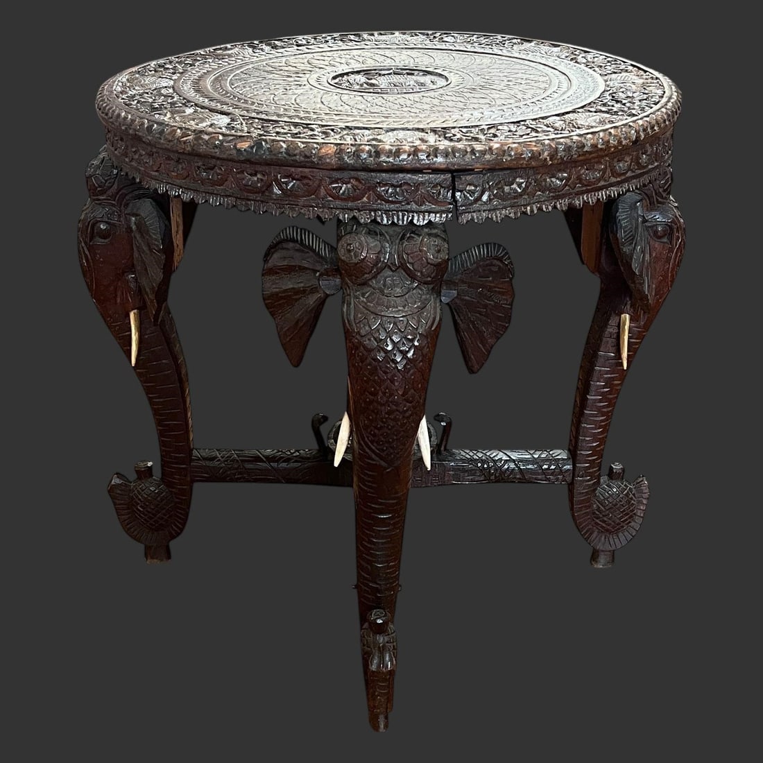 Anglo-Indian Carved Wooden Elephant Table: The table features three legs intricately carved in the form of elephant heads and trunks. The elephants are complete with bone tusks. The circular top and apron (skirt) feature extensive, detailed ha