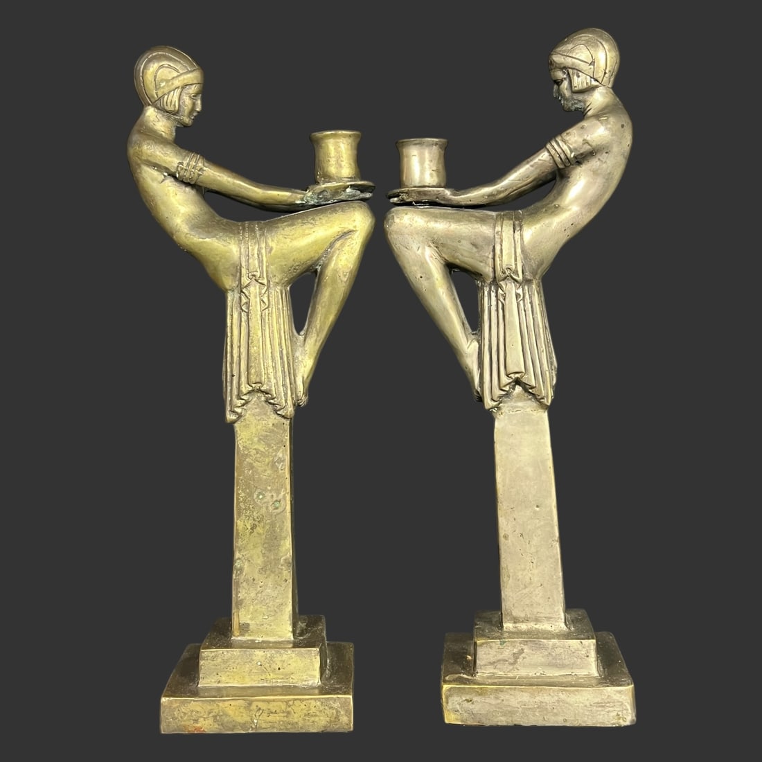 A Pair of Art Deco Style Metal Candlestick Holders: Striking pair of Art Deco figural candlesticks depicting stylized seated female forms, cast in metal with a warm natural patina. Each figure is modeled with elongated limbs and geometric drapery, hold
