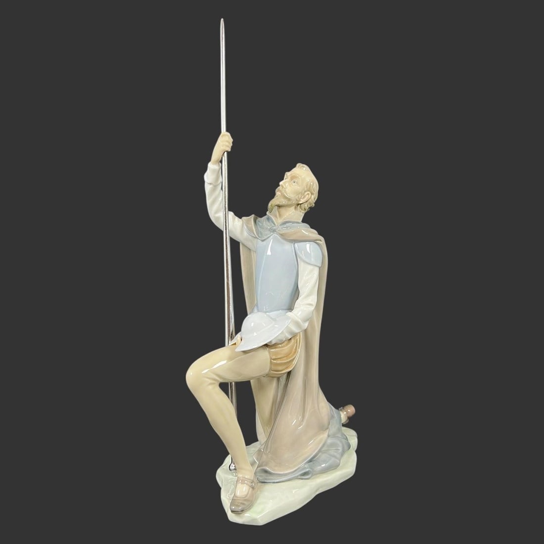Lladro Spain Porcelain #5224 Don Quixote The Quest: This elegant glazed porcelain figurine by Lladro depicts a romanticized medieval knight kneeling on one knee while raising a long lance skyward, his helmet held respectfully in his opposite hand. The