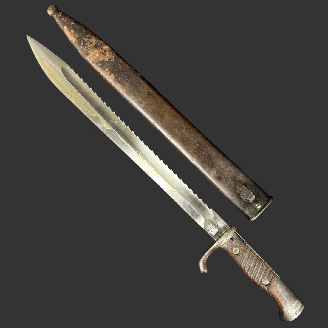 WWI German Sawback Bayonet Knife by J.H. Becker Solingen w/ Scabbard: An original German World War I–era sawback bayonet, featuring a long, single-edged blade with pronounced spine serrations and a distinctive hooked quillon. The ricasso is clearly stamped “J.H. Bec