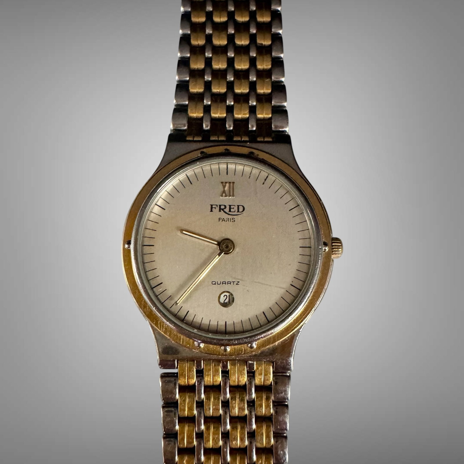 Vintage FRED Paris Quartz Wristwatch – Two Tone Bracelet – Swiss Dress Watch: Elegant vintage FRED Paris quartz wristwatch featuring a minimalist champagne dial with Roman numeral XII marker, date window at 6 o’clock, and integrated two-tone bracelet. A refined French lux