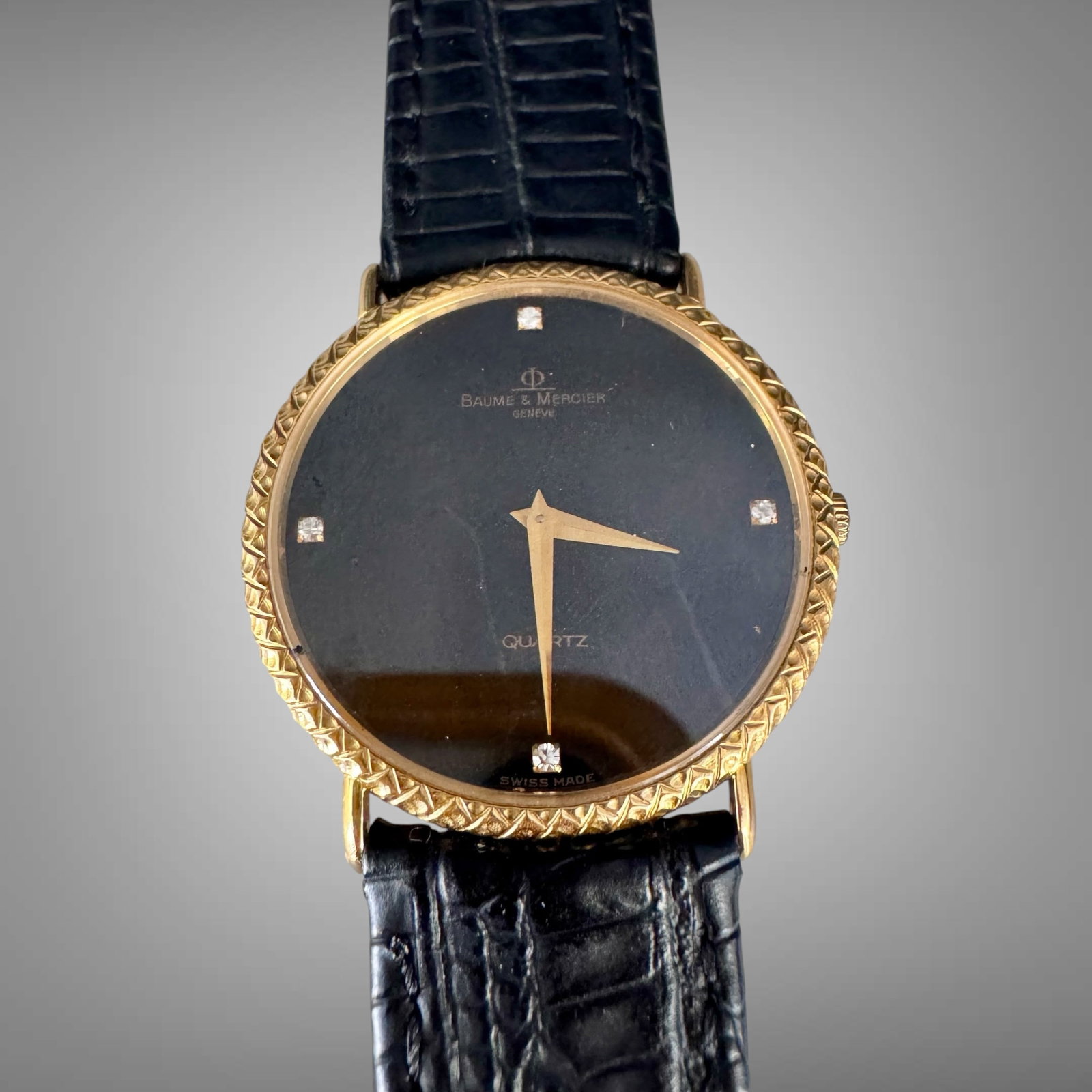 Baume & Mercier 18K Gold Quartz Watch Black Dial 082: Elegant Baume & Mercier Genève quartz wristwatch featuring a black dial with diamond-style hour markers, gold-tone hands, and a textured 18K gold case. The watch is paired with a black liza
