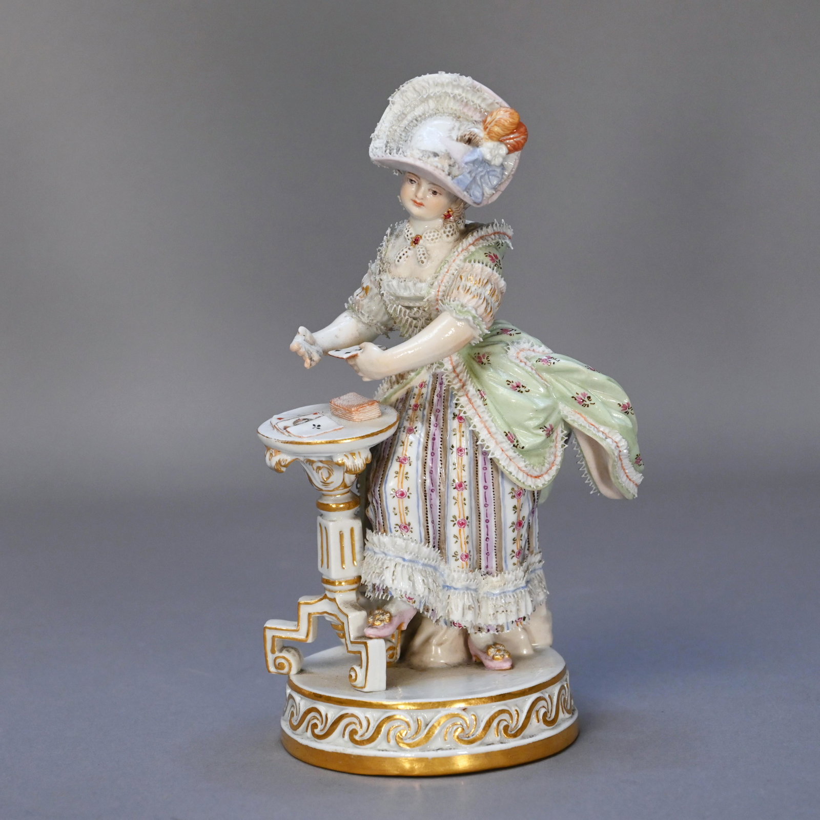 Meissen Porcelain Figure of a Woman Playing Cards: Modeled as a standing female figure with foot raised upon the leg of a stand upon which she is dealing playing cards, raised on a circular plinth base with gilt decorated scrolled edge. H: 6.25 in., W