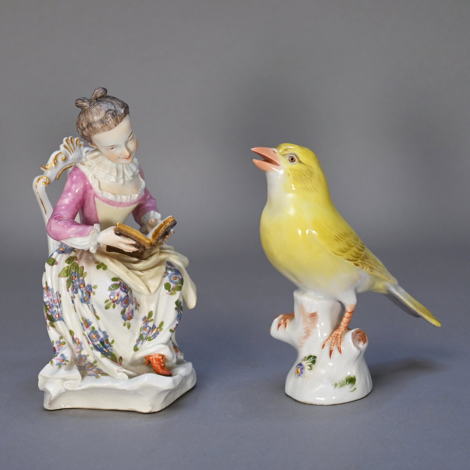 Two Meissen Porcelain Figures: The first, a model depicting a seated woman reading with book in her lap; the second a model of a yellow song bird perched on a stump. H: 5.25 in., W: 3 in., D: 3 in. (first) H: 4.25 in., W: 1.5 in., 