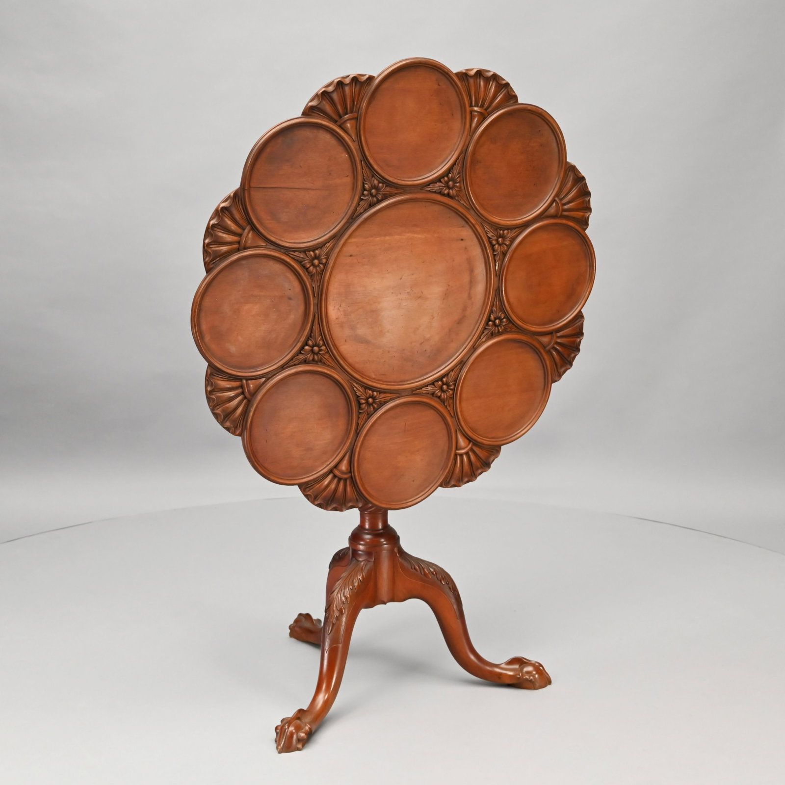 English Carved Mahogany Tilt-top Dessert Table: The circular lobed top with central molded recession surrounded by eight smaller recessions, all divided by foliage and shells, the underside with affixed battens and pivoting on a block support, abov