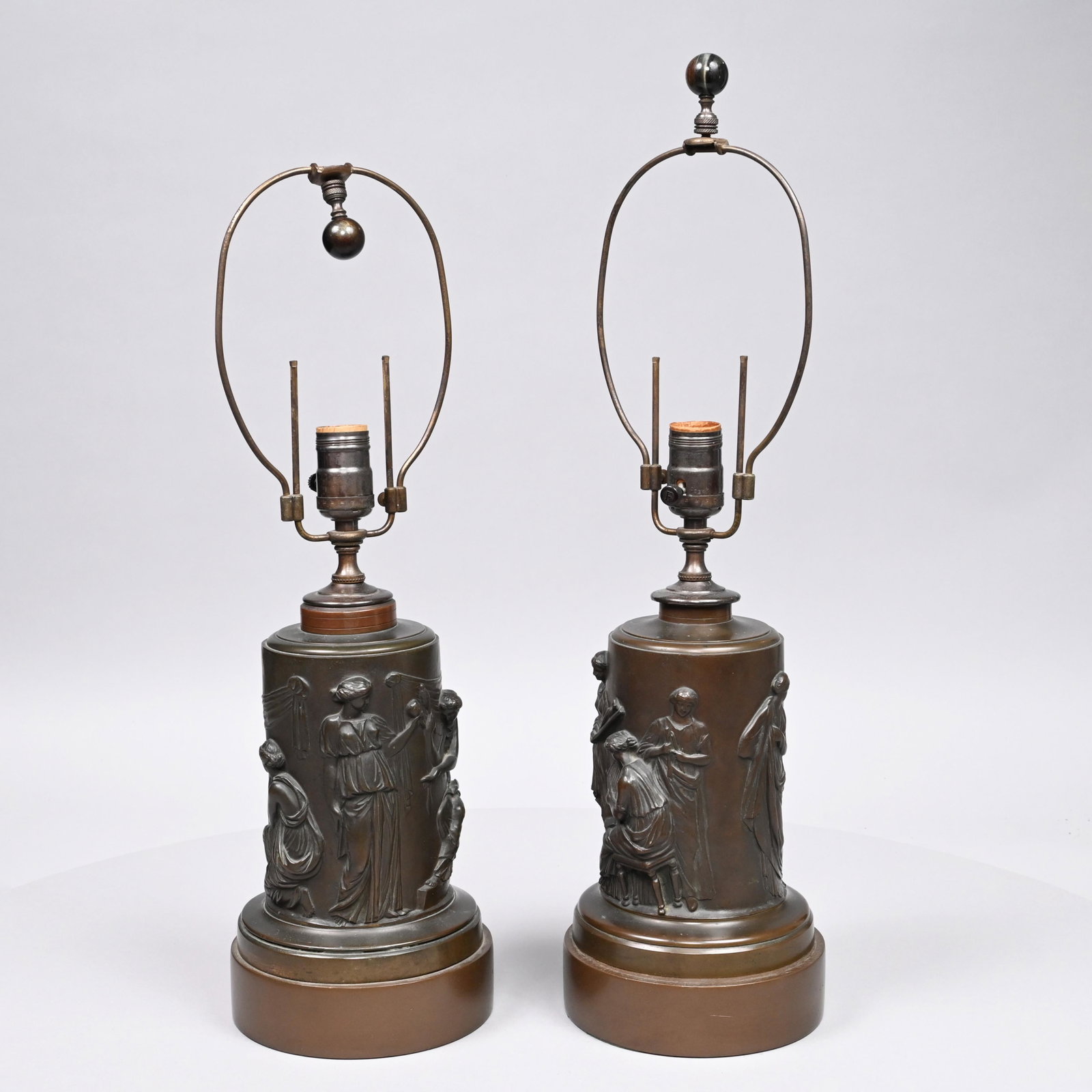 Pair of Neoclassical Style Patinated-Metal Lamps: Of cylindrical form, each cast in high relief around the sides with robed muses engaged in various activities. Height to top of harp: 20.5 in., D: 6.375 in.