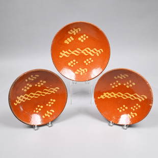 Three Pennsylvania Redware Slip-Decorated Plates: Nineteenth Century. Each painted in yellow slip across the center with three entwined wavy lines, within three groups of six dots, within a coggled rim edge. Provenance: This lot accompanied by a