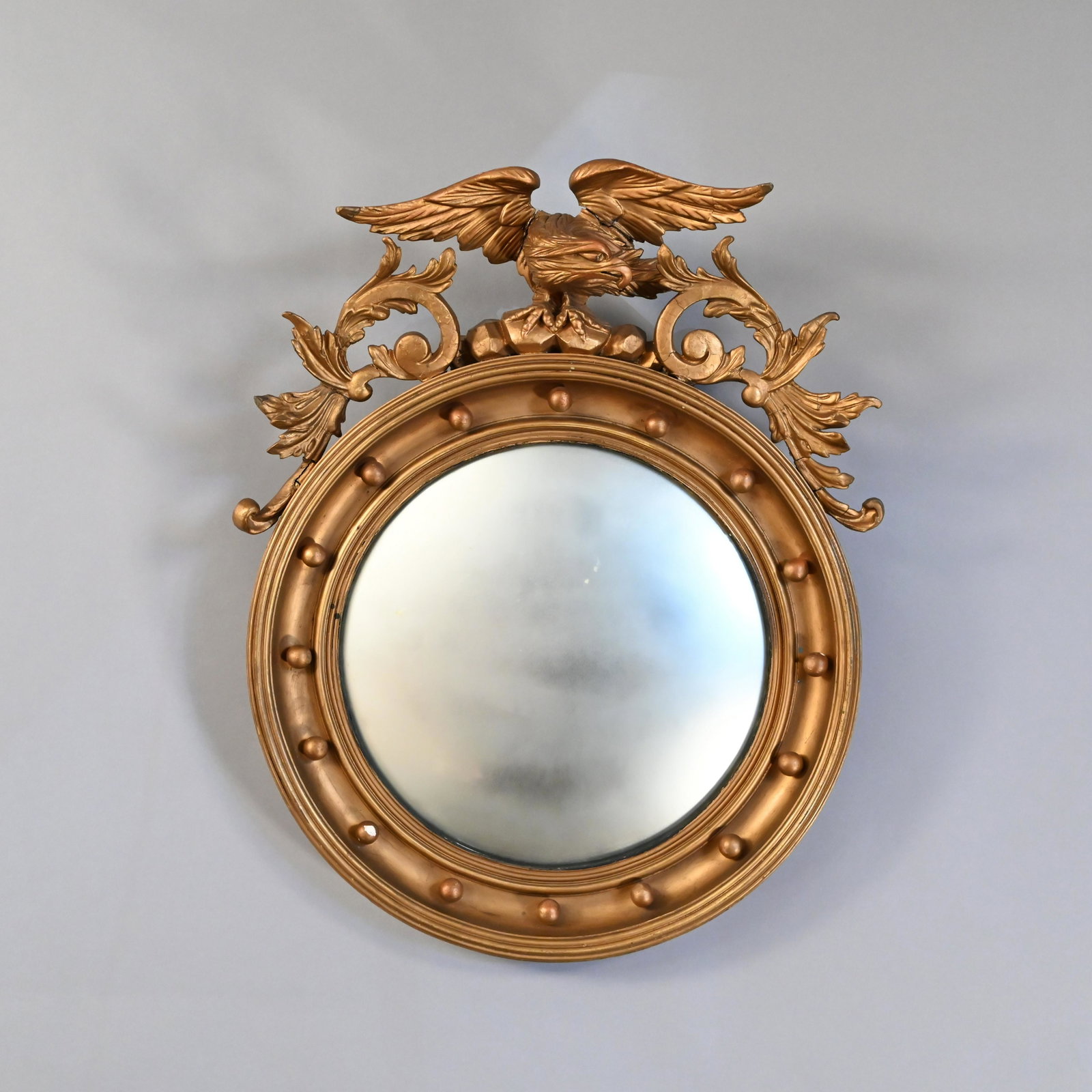 English Giltwood Convex Mirror: The circular mirror plate within a cove-molded frame affixed with spherules, surmounted by a carved displayed eagle perched on a rocky outcropping and flanked by trailing vinery. H: 28 in., W: 24 in.,