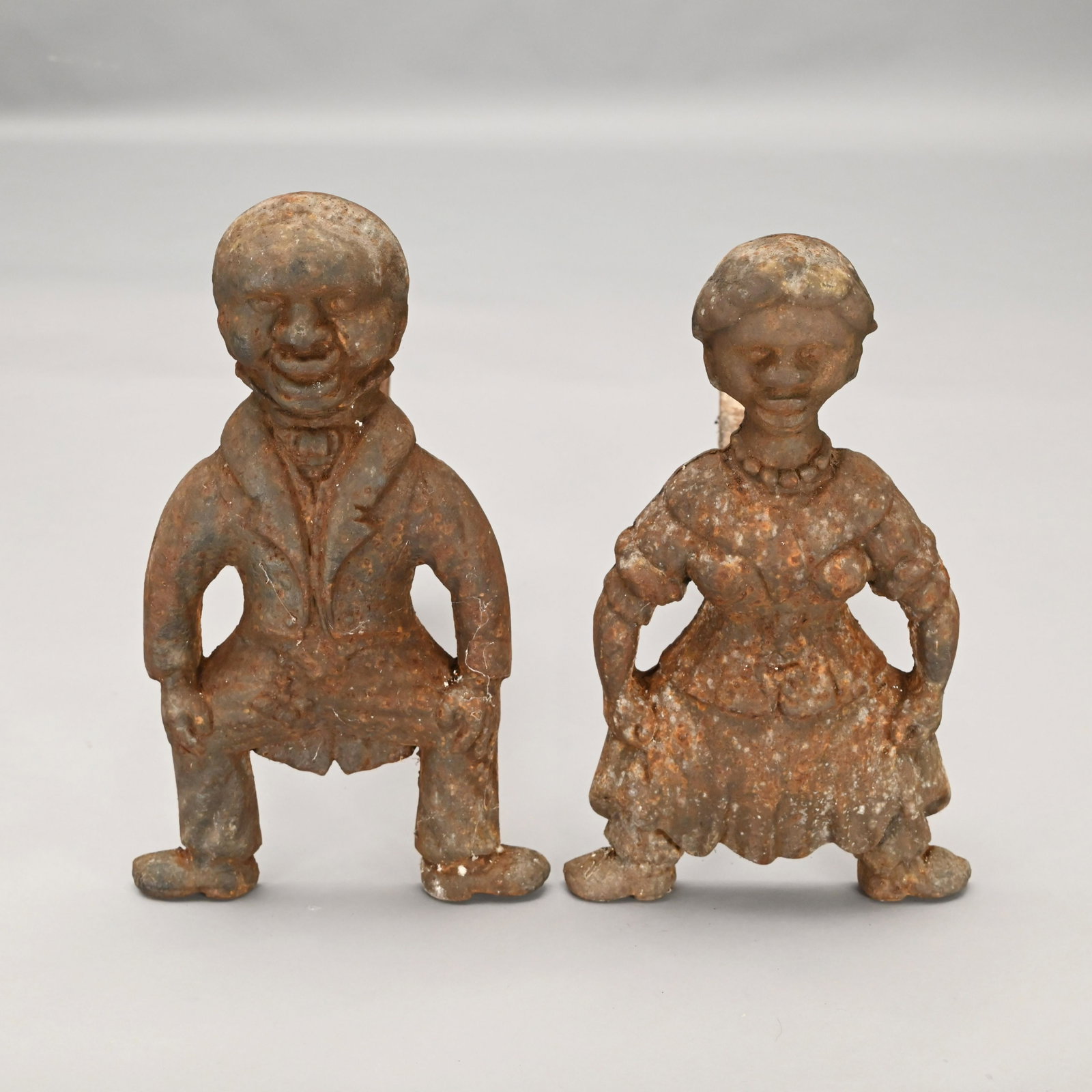 Pair of American Figural Cast Iron Andirons: Caricaturesque depiction of an African American couple, the gentleman in a suit with tails and the lady wearing a dress with necklace and stole. Each with pistol form dogs. H: 17 in., W: 10 in., D: 18