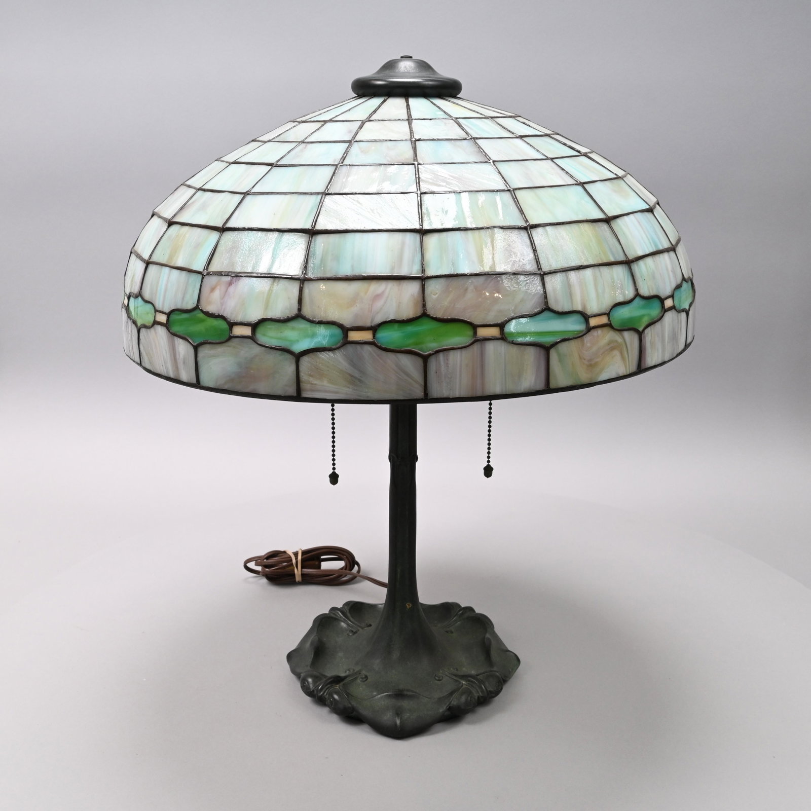 American Leaded Glass & Patinated-Metal Table Lamp: J.A. Whaley for McKenney & Waterbury, Boston, Massachusetts, Circa 1910. The tile shade surrounded by a border of green shaped lozenges, the patinated-bronze base cast with lobed foliate devices, clot