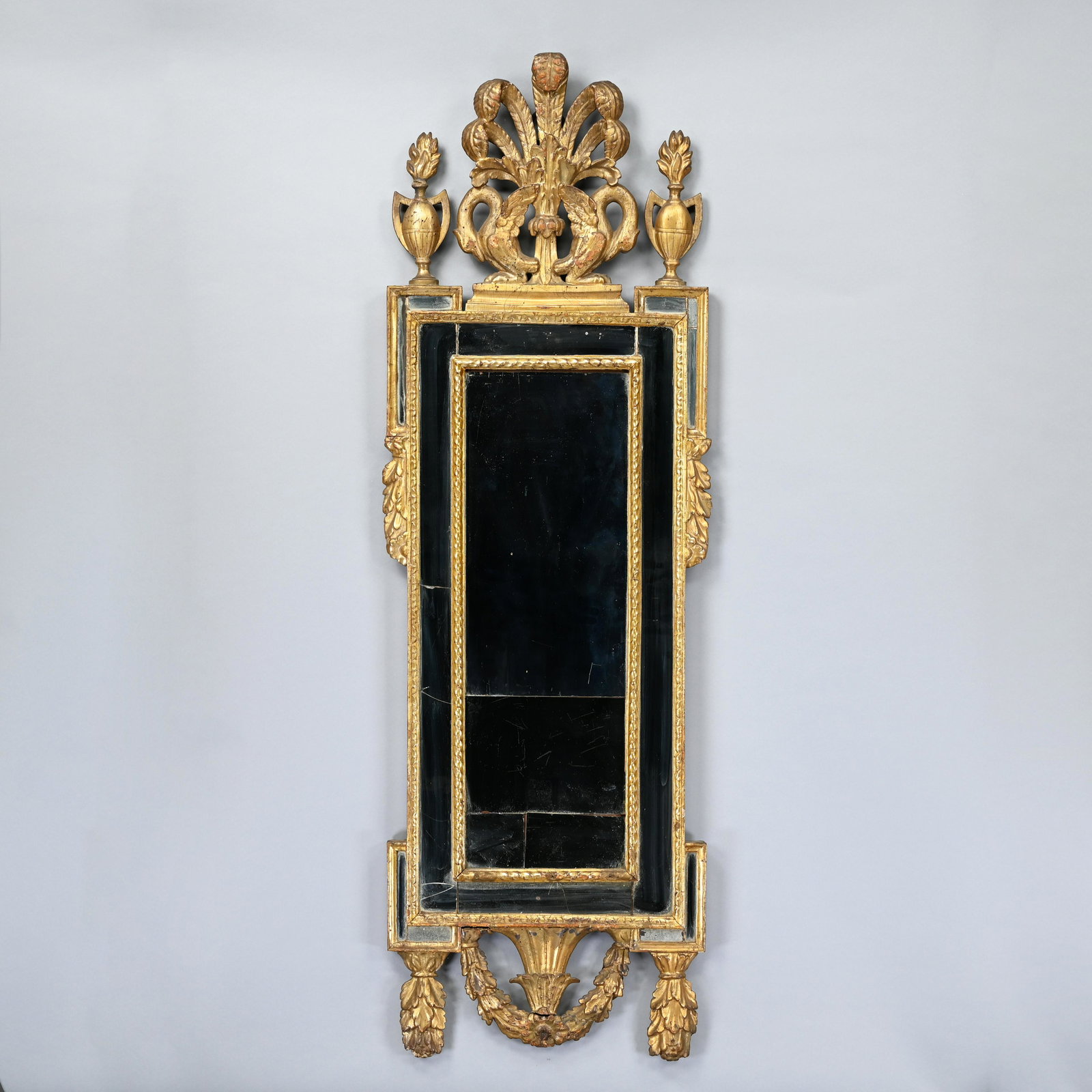 Continental Neoclassical Carved Giltwood Mirror: The crest with feathers and two swans, flanked by urns with flame finials above a mirror plate over a conforming valance. H: 62 in., W: 19.5 in., D: 2 in.