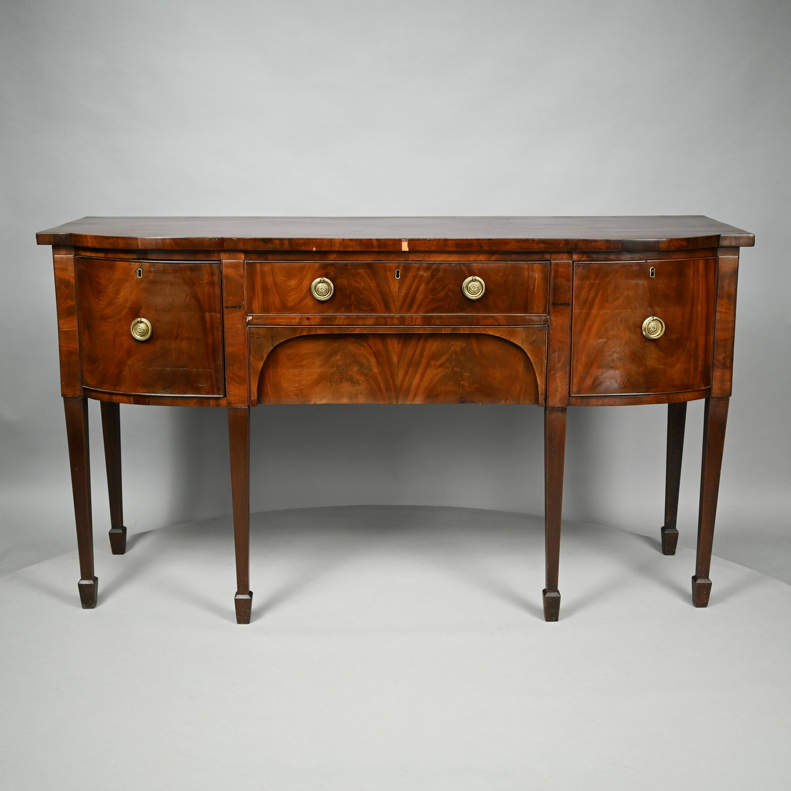 George III Mahogany Sideboard: Early Nineteenth Century. The rectangular top with bow-front above one cockbeaded drawer over an arched recessed drawer, flanked by a cupboard and a cellarette/bottle drawer, raised on square tapered 