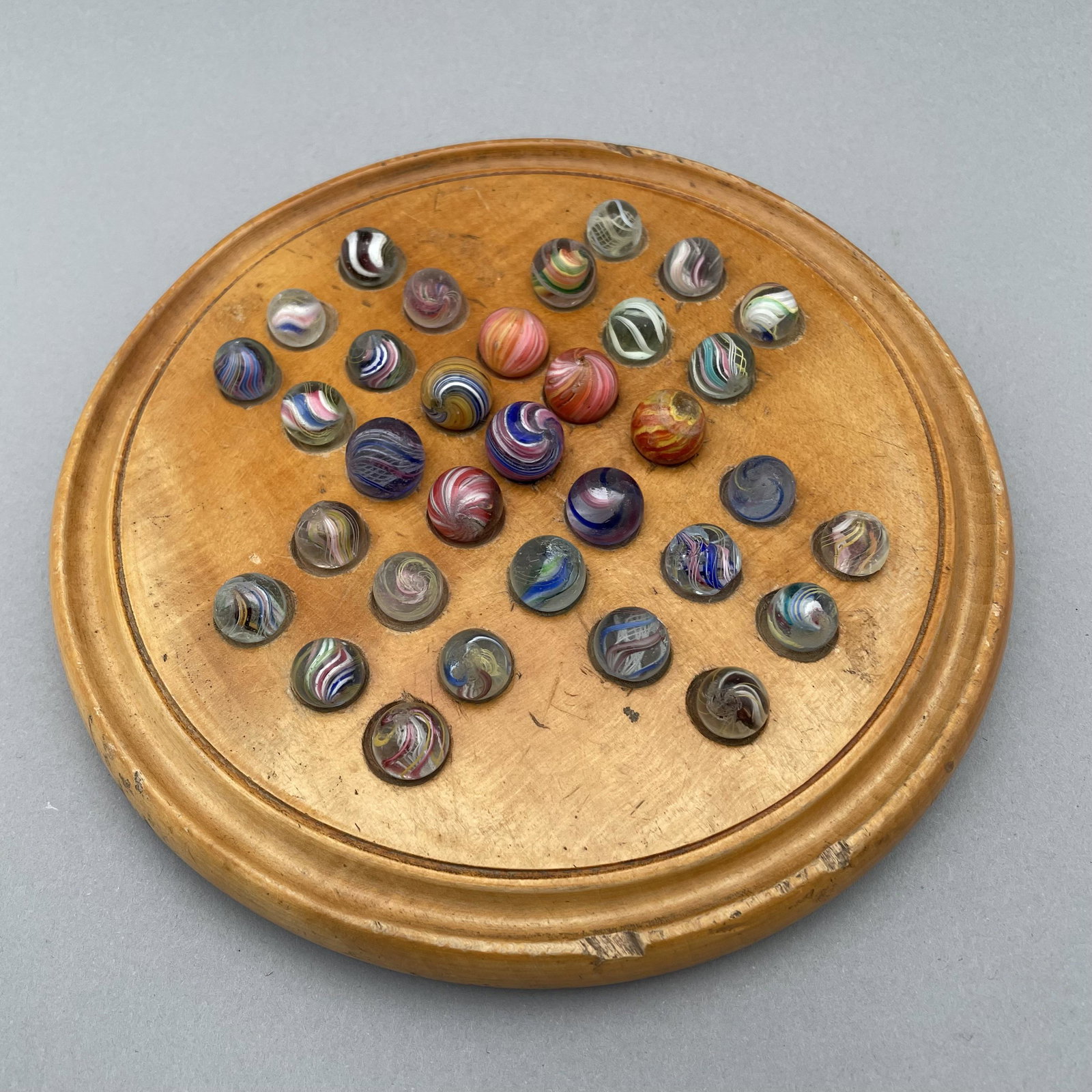 Solitaire Board with Thirty-Three Pee Wee Marbles: The marbles handmade and comprising swirls and onion skins. Approximate range of diam: 29/64 to 19/32 in. Board: Diam: 7.5 in., D: .75 in.